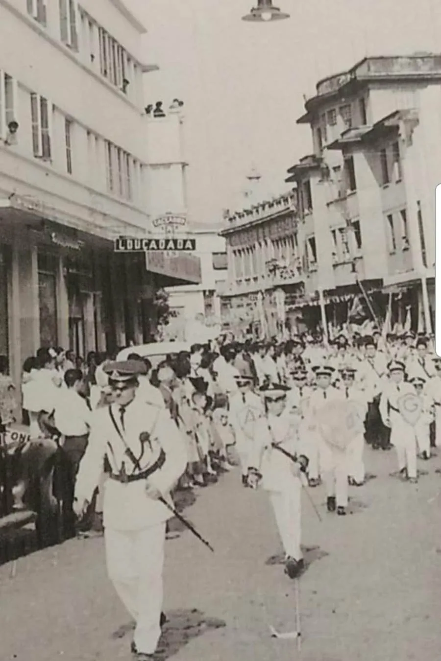 Fotografia antiga de um desfile na Rua Rui Barbosa, com o letreiro Louça Dada acima das pessoas